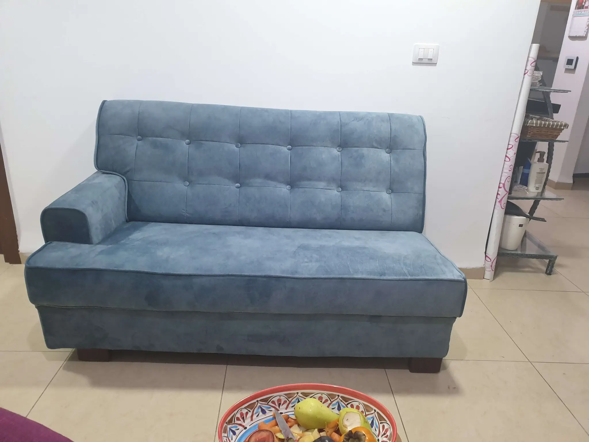 ספה כחולה בעלת משענת כפתורים מרופדת. הספה ממוקמת בחלל ביתי על ריצוף קרמי בצבע בהיר. על הרצפה מלפני הספה, יש צלחת צבעונית עם פירות. ברקע ניתן לראות קיר לבן עם מתגי חשמל ומדף קטן בצד ימין